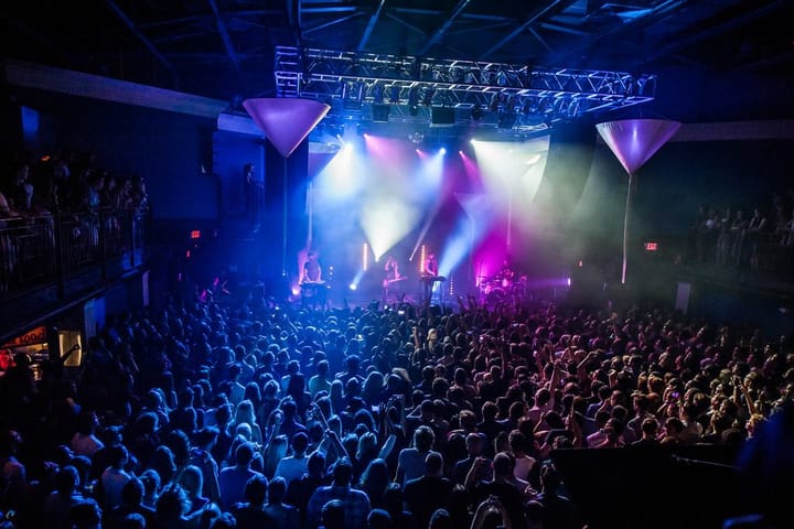 A crowd stands before a stage lit up by several different colored lights as band members play to the audience.
